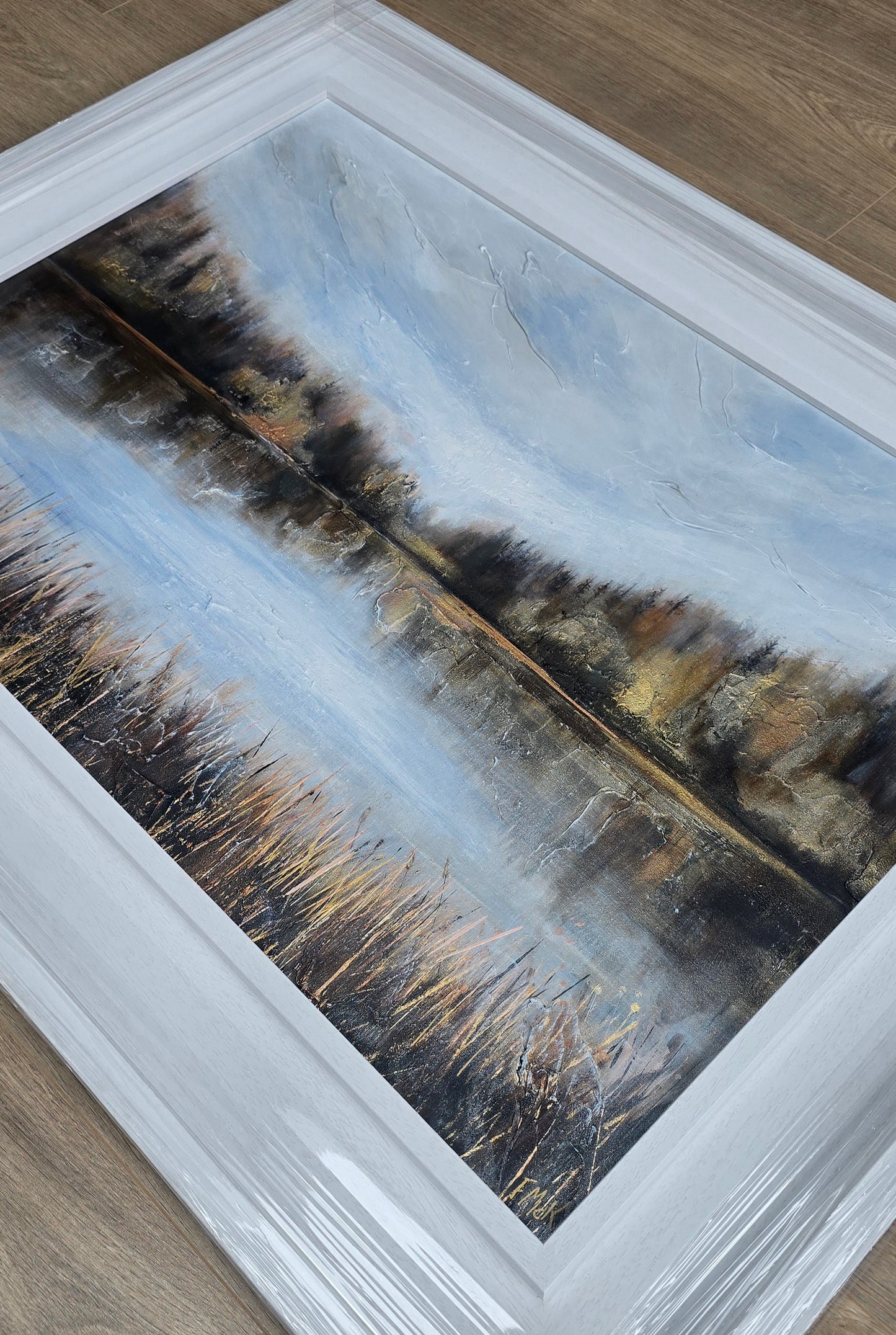 Framed artwork of a landscape with trees and water on a wooden floor.