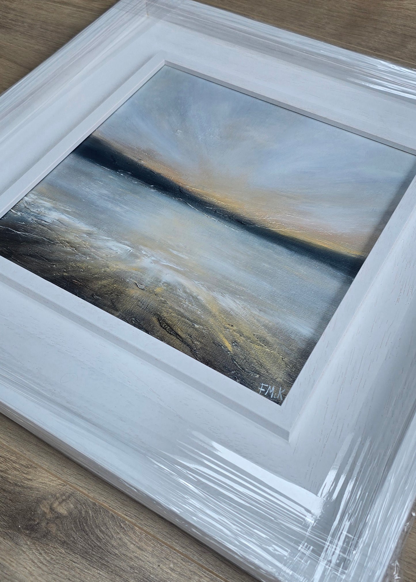 Abstract sunset painting in a white frame on a wooden surface