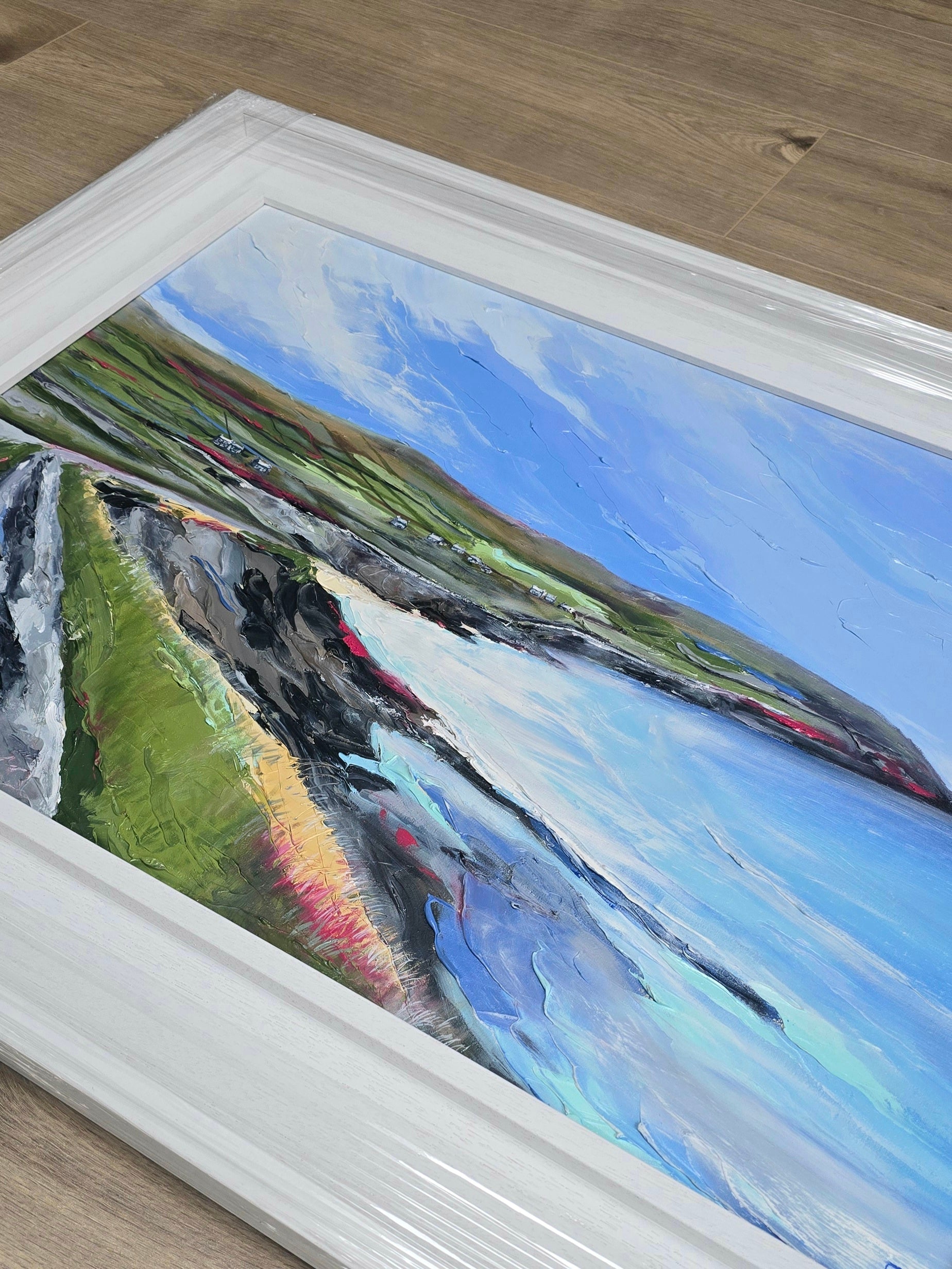 Framed painting of a landscape with a river and mountains on a wooden floor.