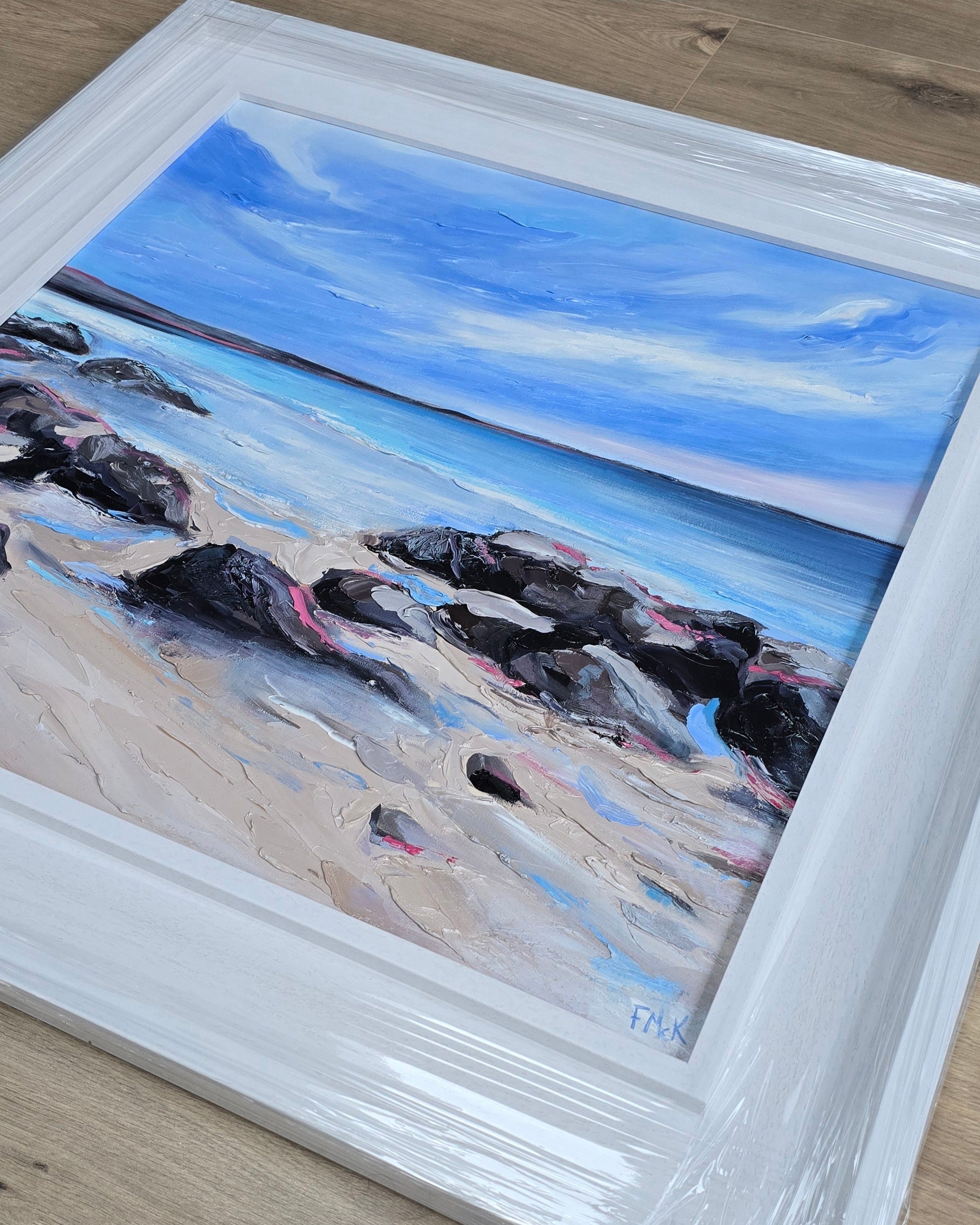 Framed painting of a beach scene with rocks and blue sky.