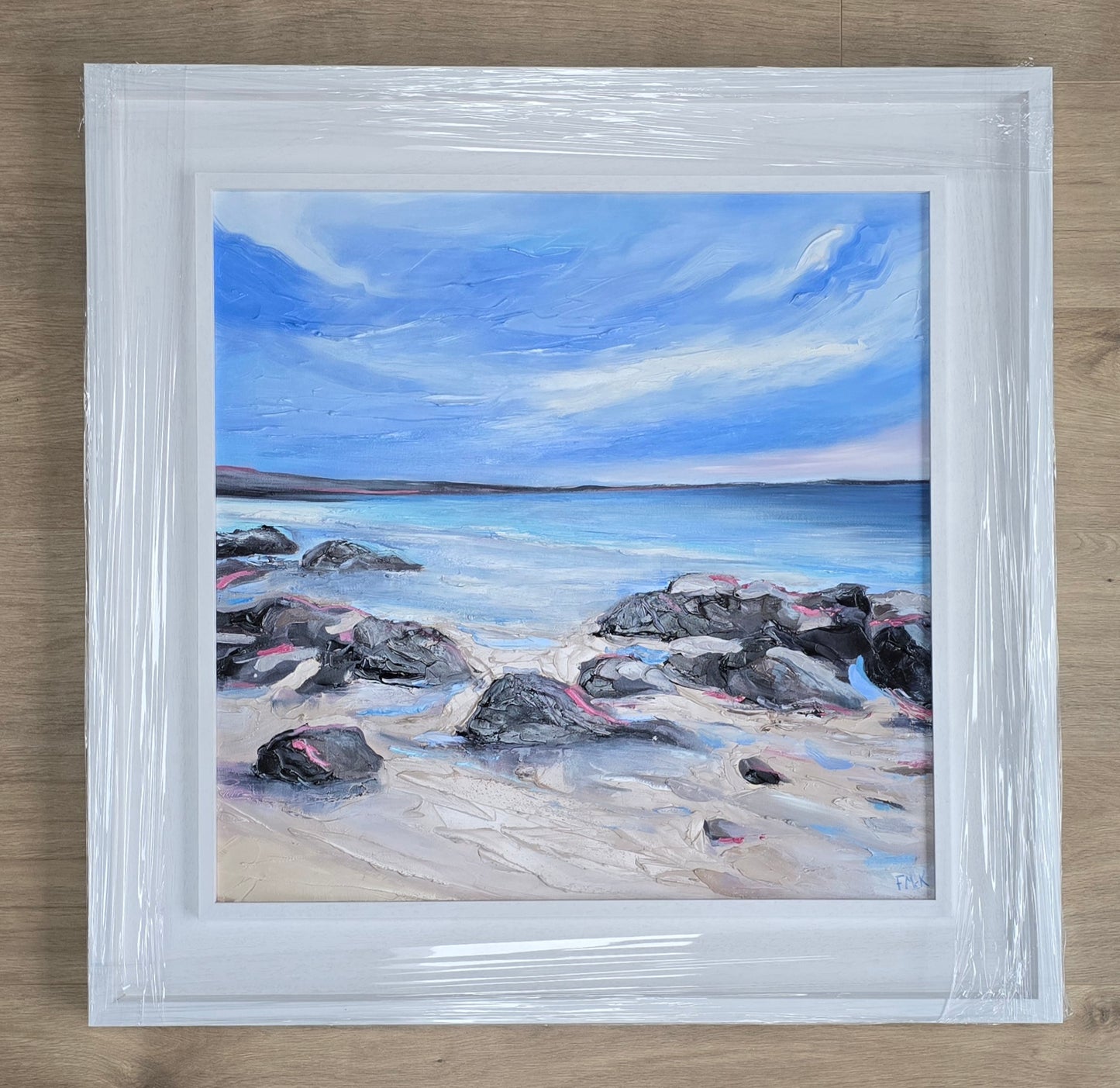 Framed painting of a beach scene with rocks and blue sky.