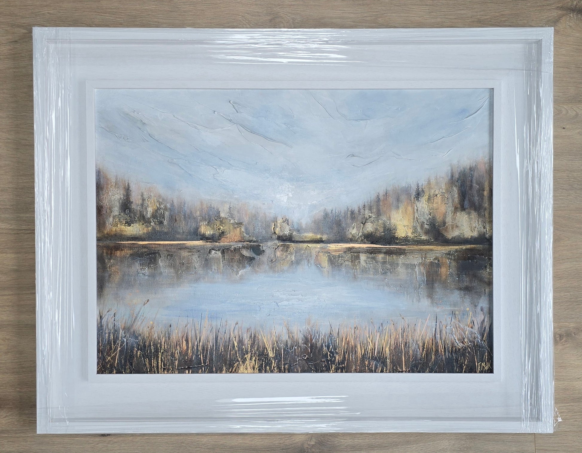 Framed painting of a landscape with a lake and trees on a wooden surface