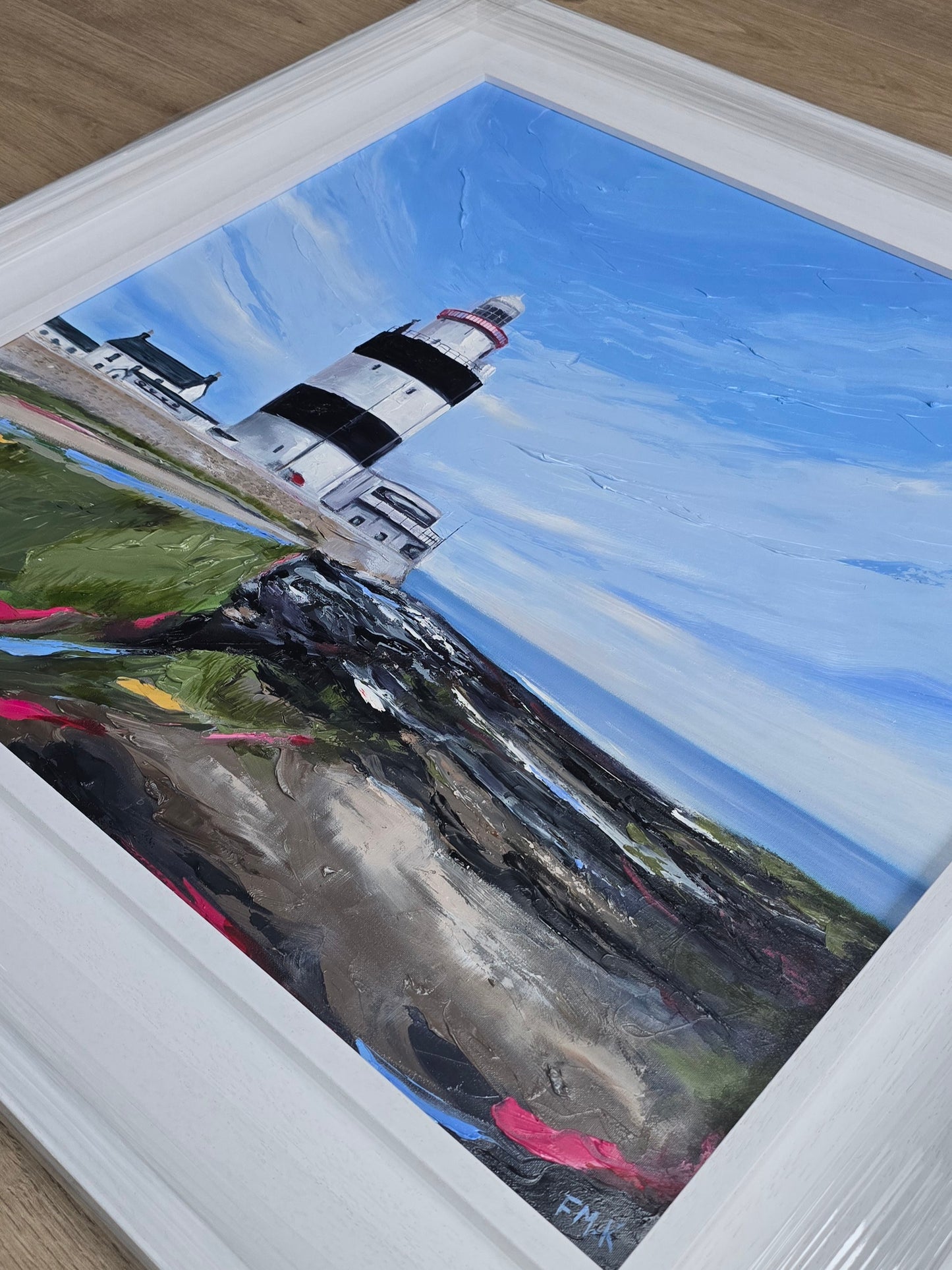 Framed painting of a lighthouse on a hillside with a blue sky.