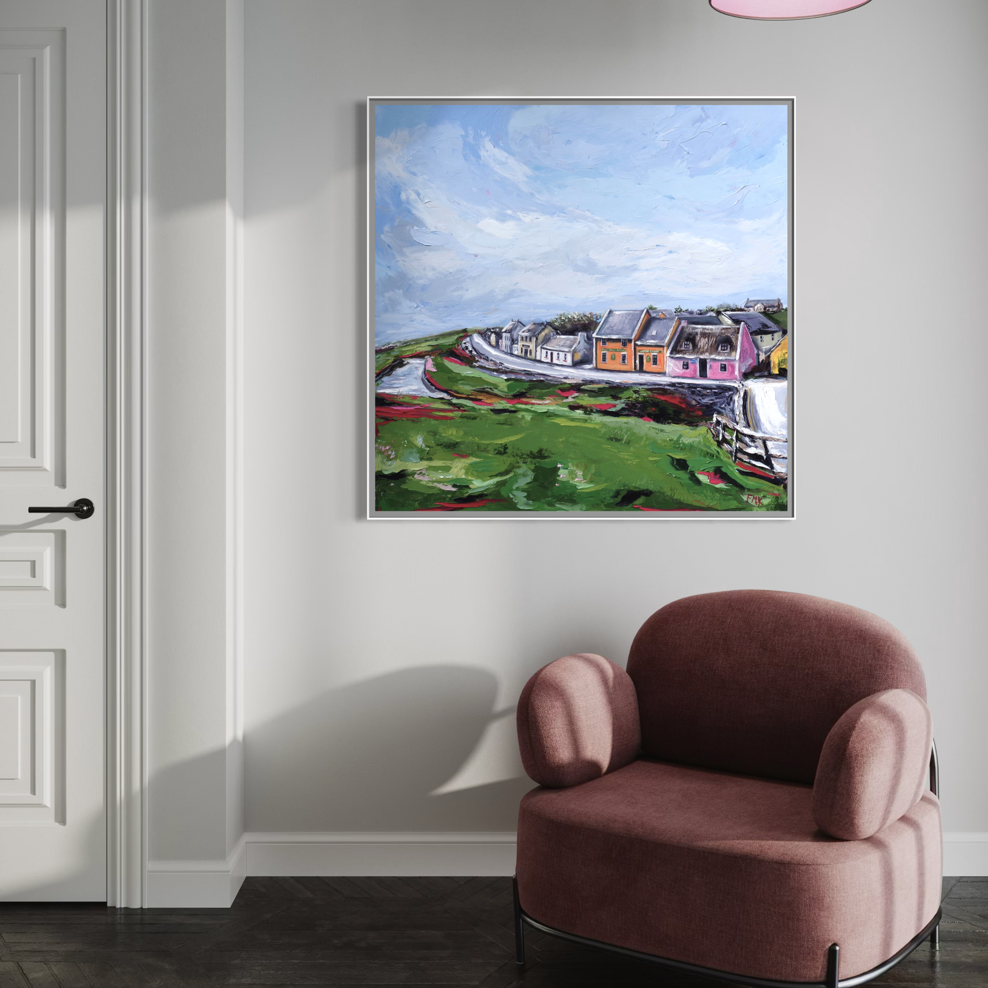 Framed painting of a coastal village scene on a wall above a pink armchair.