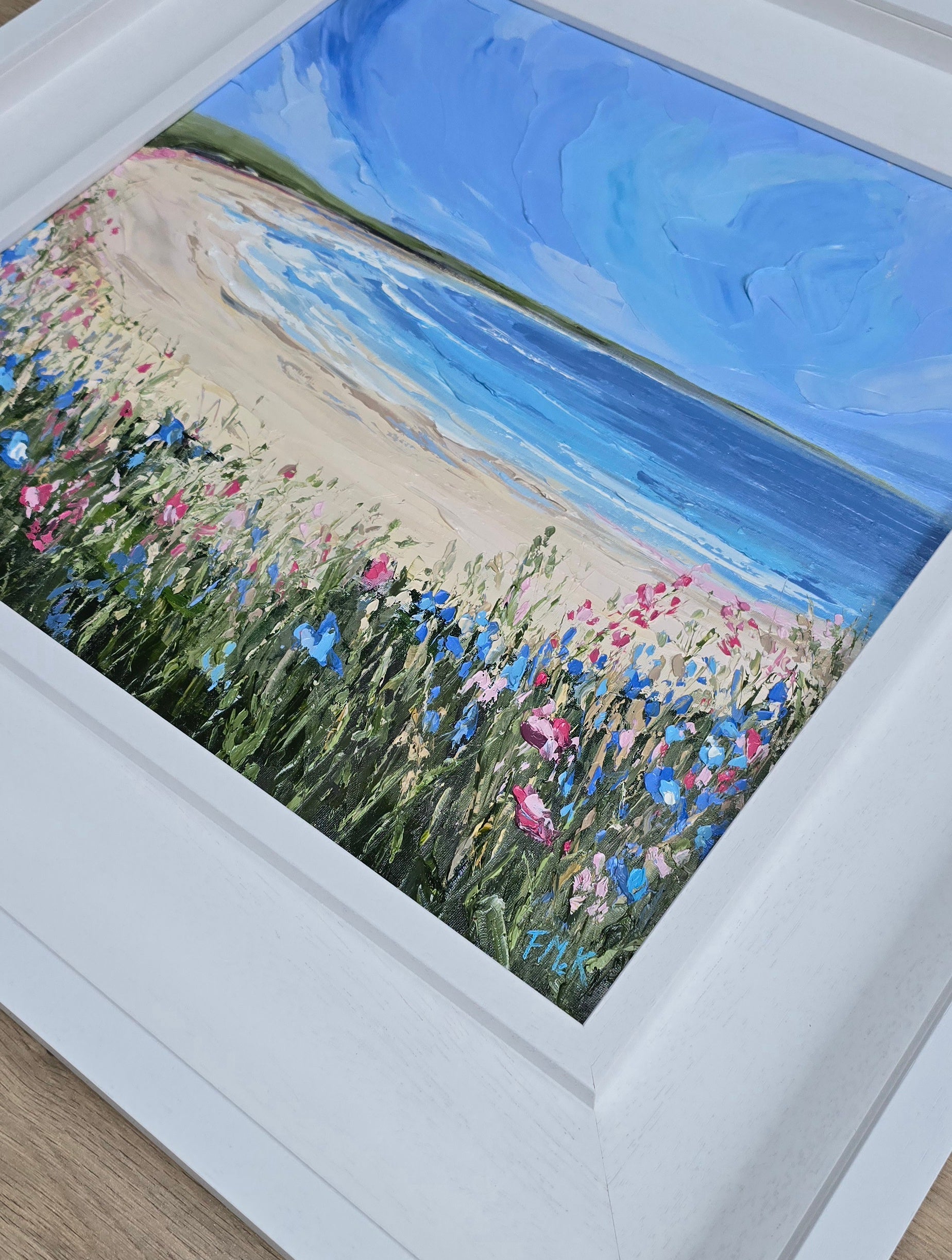 Framed artwork of a beach scene with flowers and blue sky.