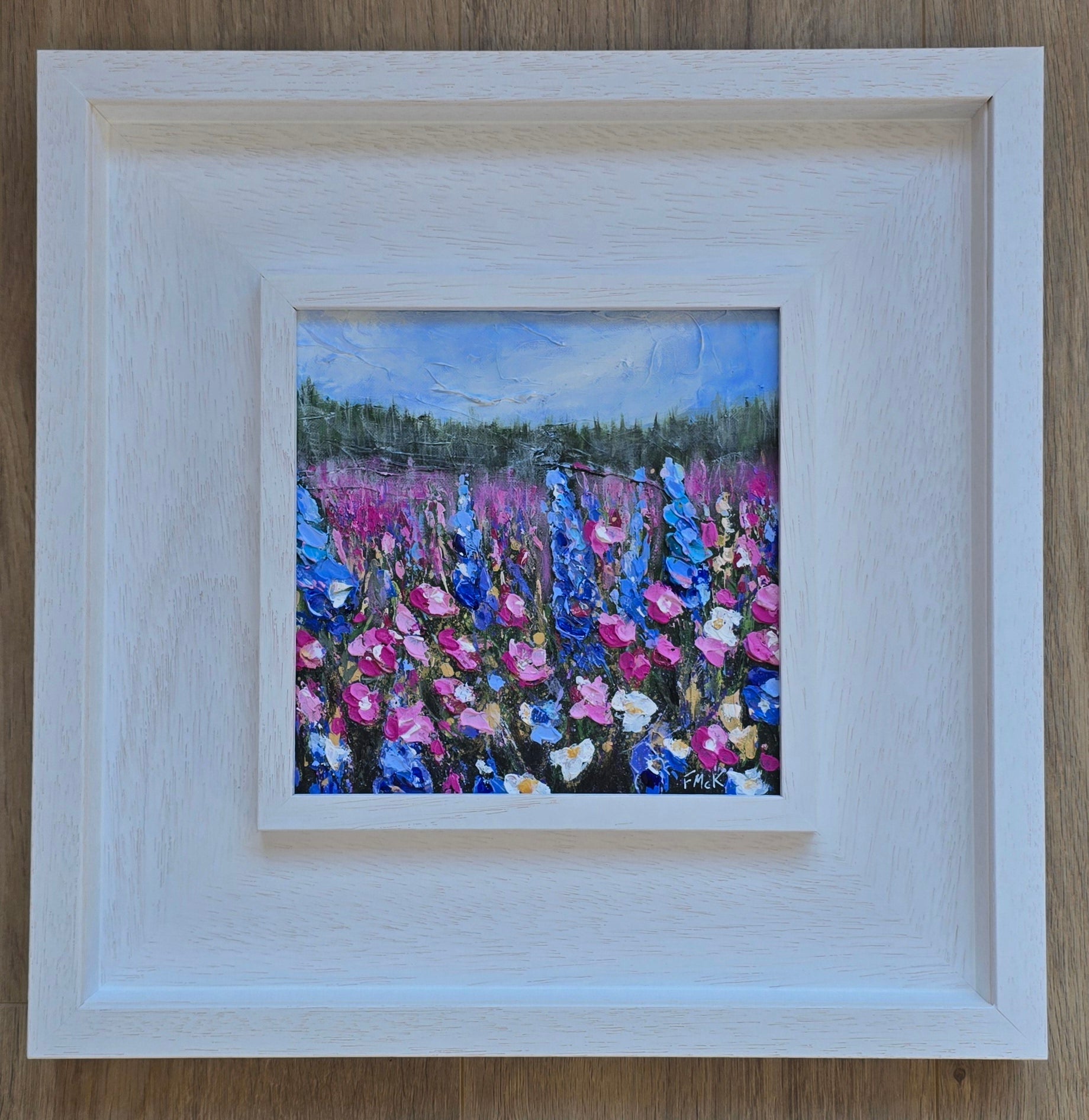 A framed acrylic painting of a field with vibrant flowers, predominantly blue and pink, with a bright sky.