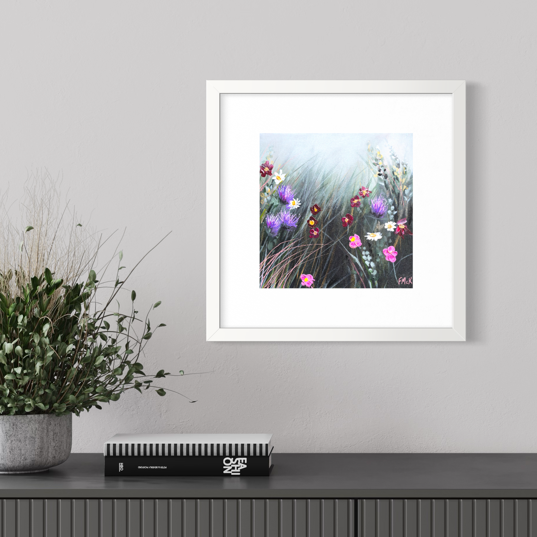 Framed artwork of flowers on a wall above a shelf with a plant and book.
